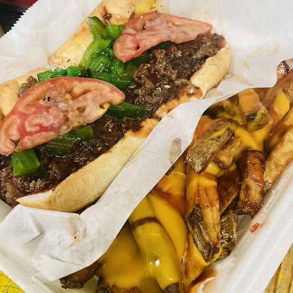 A steak hoagy with green peppers and tomatoes, alongside cheesy fries, in a white paper tray.